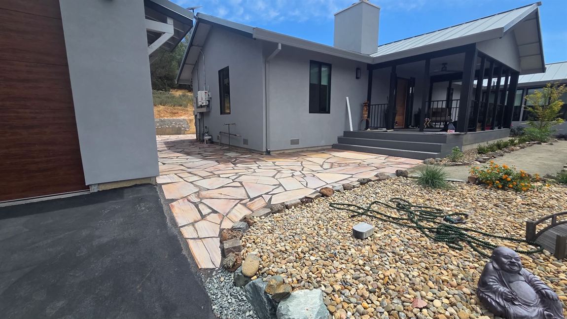 Curving flagstone walkway sweeping from a driveway toward a covered entry, with a cobble and river-rock bed and new shrubs alongside
