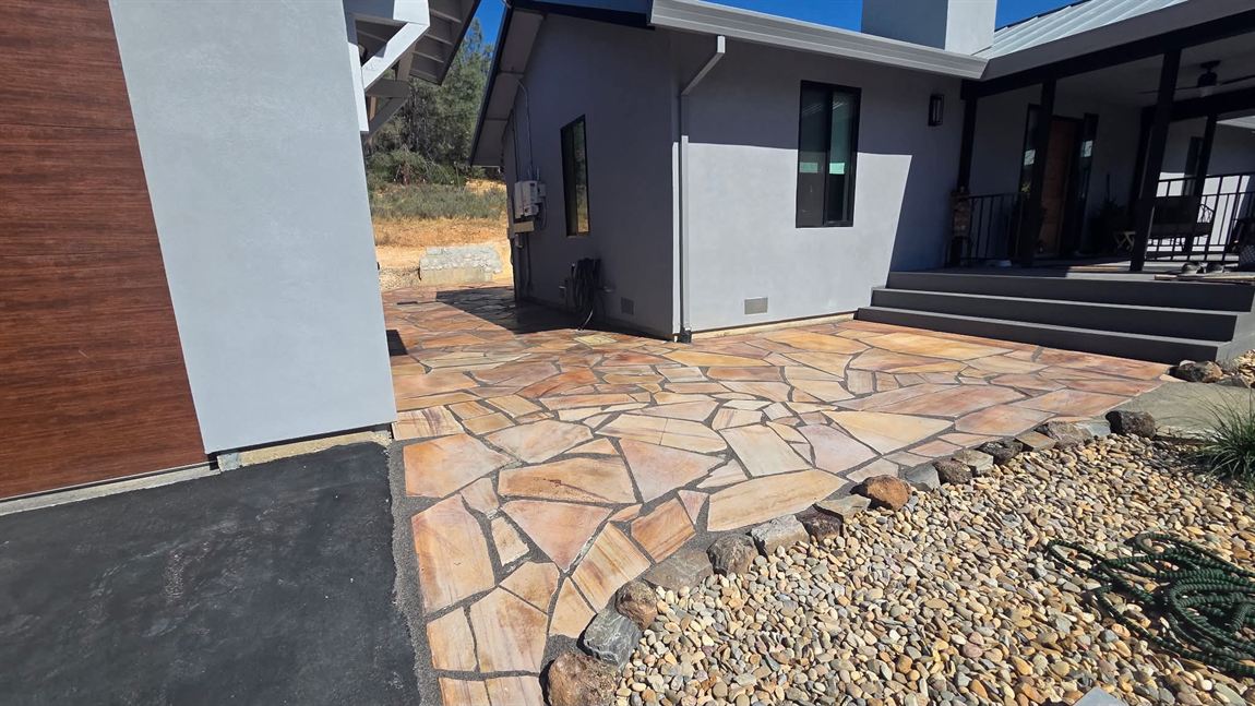 Broad irregular flagstone courtyard in warm tan and peach stone with dark mortar joints, edged by a river-rock gravel bed beside a grey stucco house